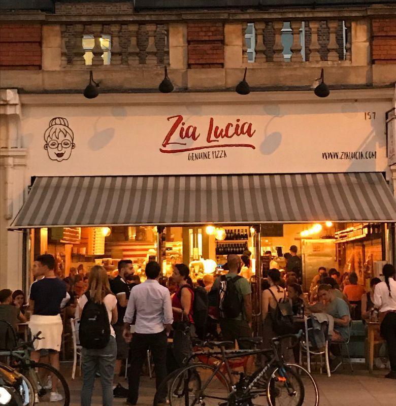 A Zia Lucia pizzeria at night, lit up under its striped awning, with a crowd outside